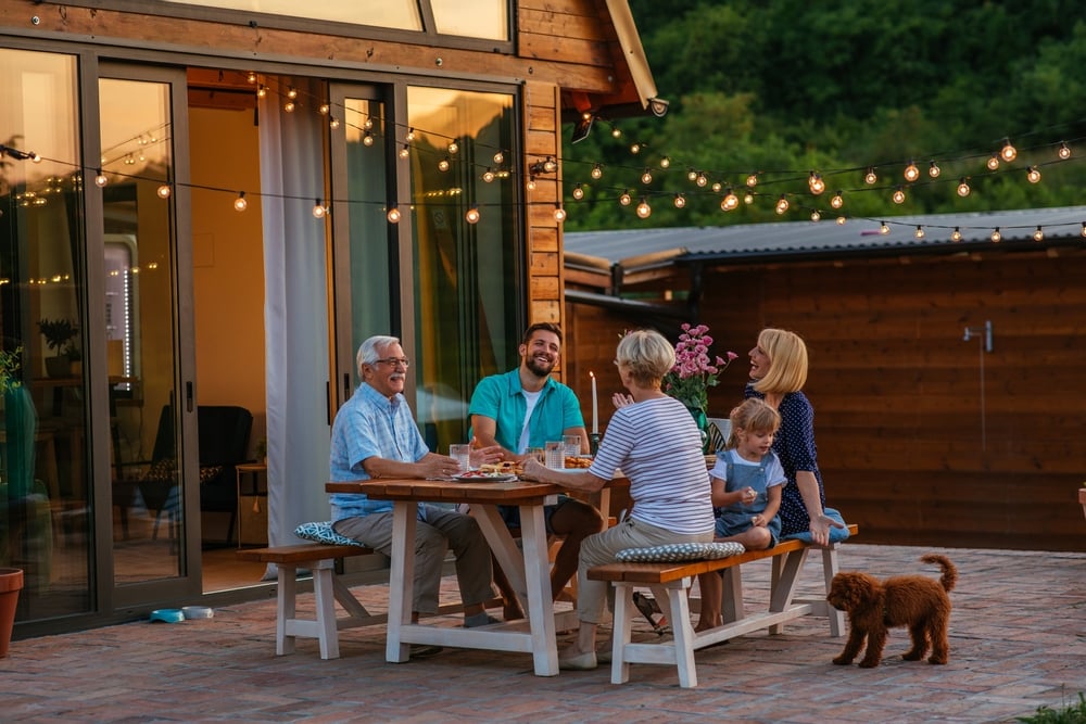 Happy,Family,Eating,Together,Outdoors.,Smiling,Generation,Family,Sitting,At A multiGenerational family enjoys a backyard dinner at a wooden patio table under string lights, with a small brown dog nearby.