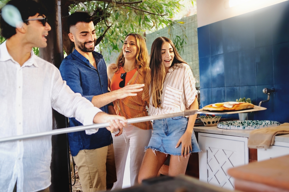 Group,Of,Friends,Have,Fun,Preparing,Pizzas,And,Baking,Them, Four friends at an outdoor kitchen laugh as one passes a pizza on a long pizza peel toward the group.