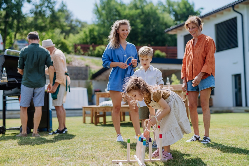 Multi,Generation,Family,Playing,Throw,A,Ring,Game,When,Grilling Family outdoor gathering in a sunny backyard; a young girl bends to knock over wooden pins while adults chat and grill nearby.