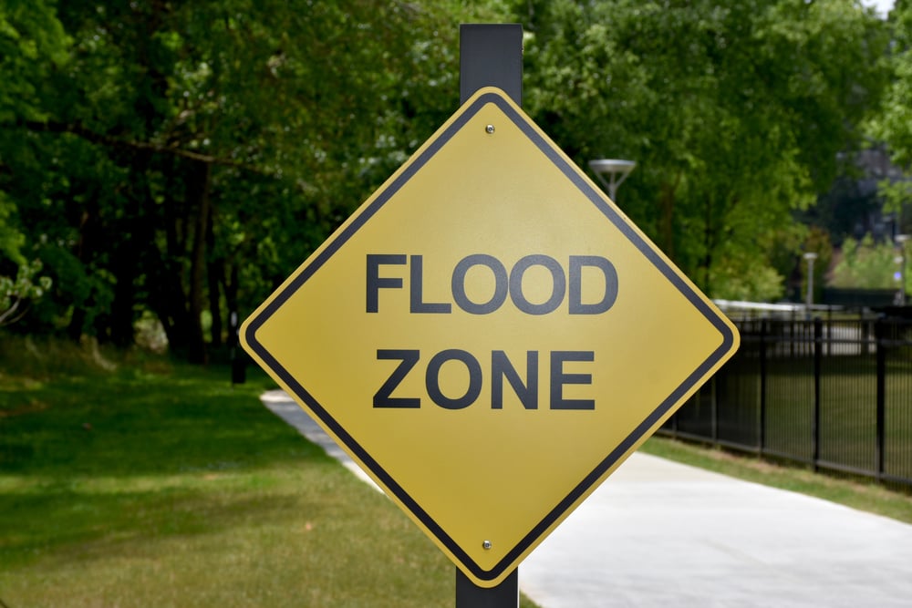 A,Flood,Zone,Warning,Sign,In,The,Park Yellow diamond flood zone sign on a post beside a paved path with trees and a fence in the background.