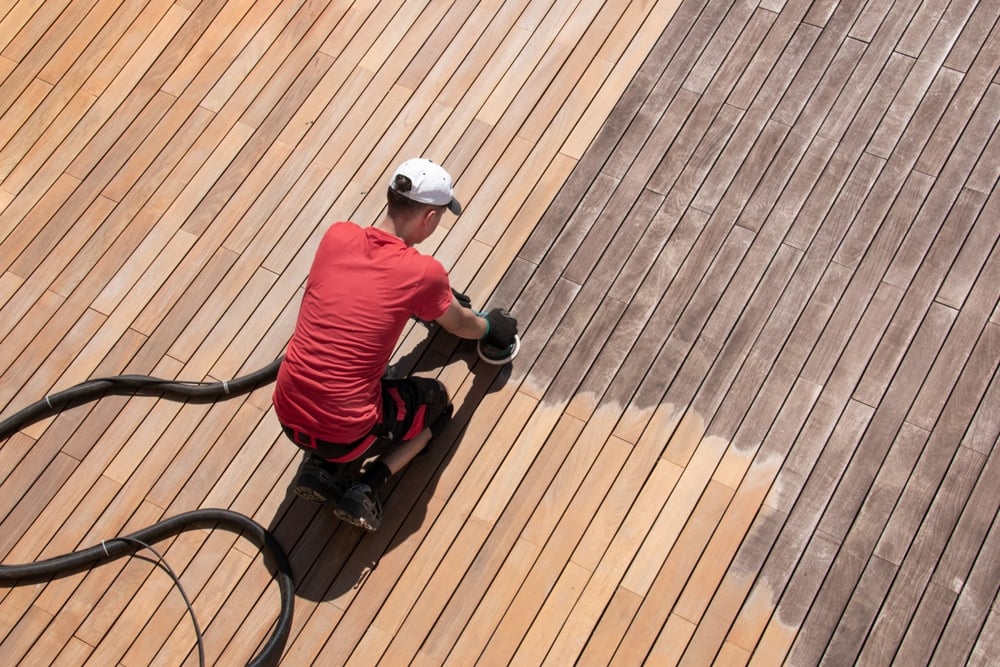 Deck,Renovation,Worker,Sanding,Ipe,Wood,Boards,Overhead,View,Space Worker in a red shirt and white cap sands a wooden deck with a power sander.