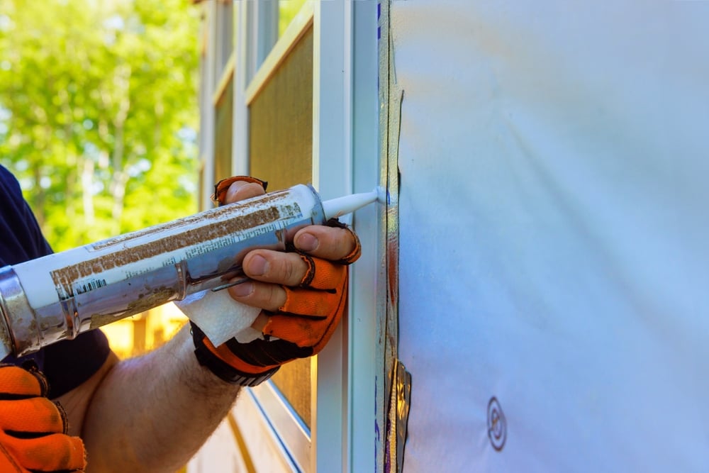 worker wearing protective gloves carefully applies caulk to edges of window frame to ensure tight seal against air leaks.